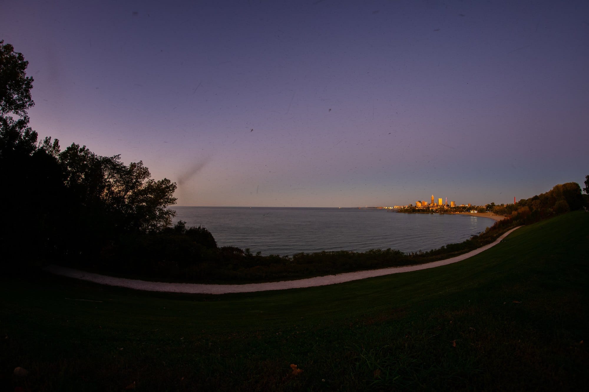A path curving across the grass above Lake Erie at dusk, with the Cleveland skyline lit on the far shore