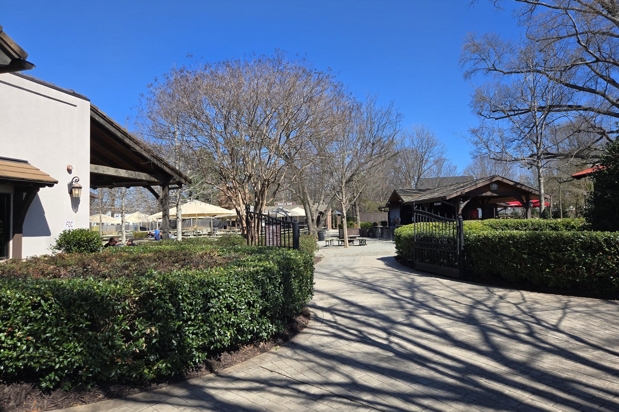 A paved path between clipped hedges leading through an open gate into a tree shaded biergarten with umbrellas and picnic tables