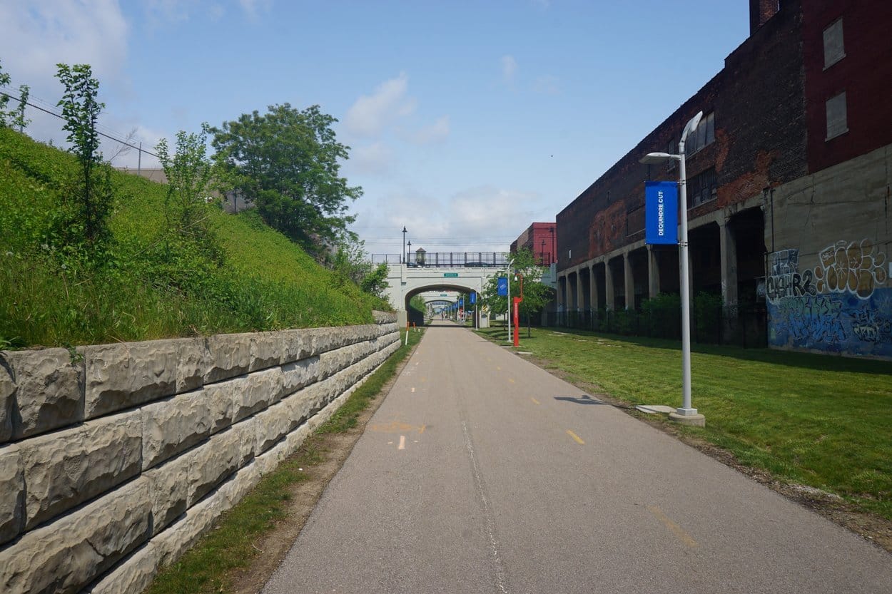 The Dequindre Cut in Detroit, a paved path below grassy banks and an old brick warehouse covered in graffiti, a stone bridge ahead
