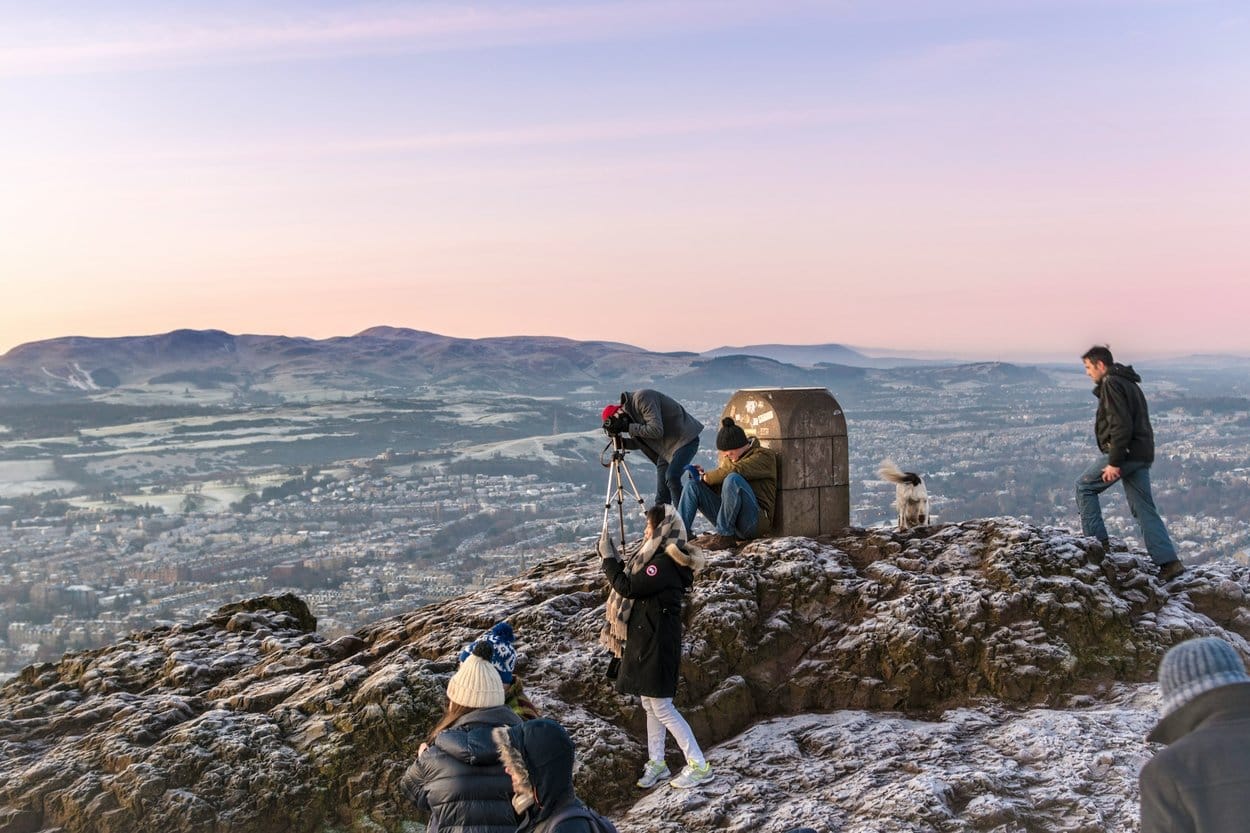 A dog and walkers on the frosty summit of Arthur's Seat at dawn, with the city and the Pentland Hills behind