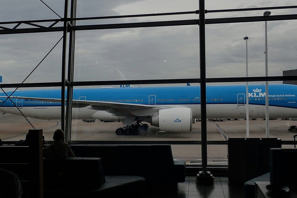 A blue KLM airliner parked at the gate, seen through a departure lounge window
