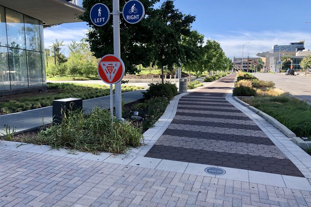 The Indianapolis Cultural Trail at Alabama and Market streets, a patterned brick path lined with grasses and young trees, with signs sending walkers left and bikes right