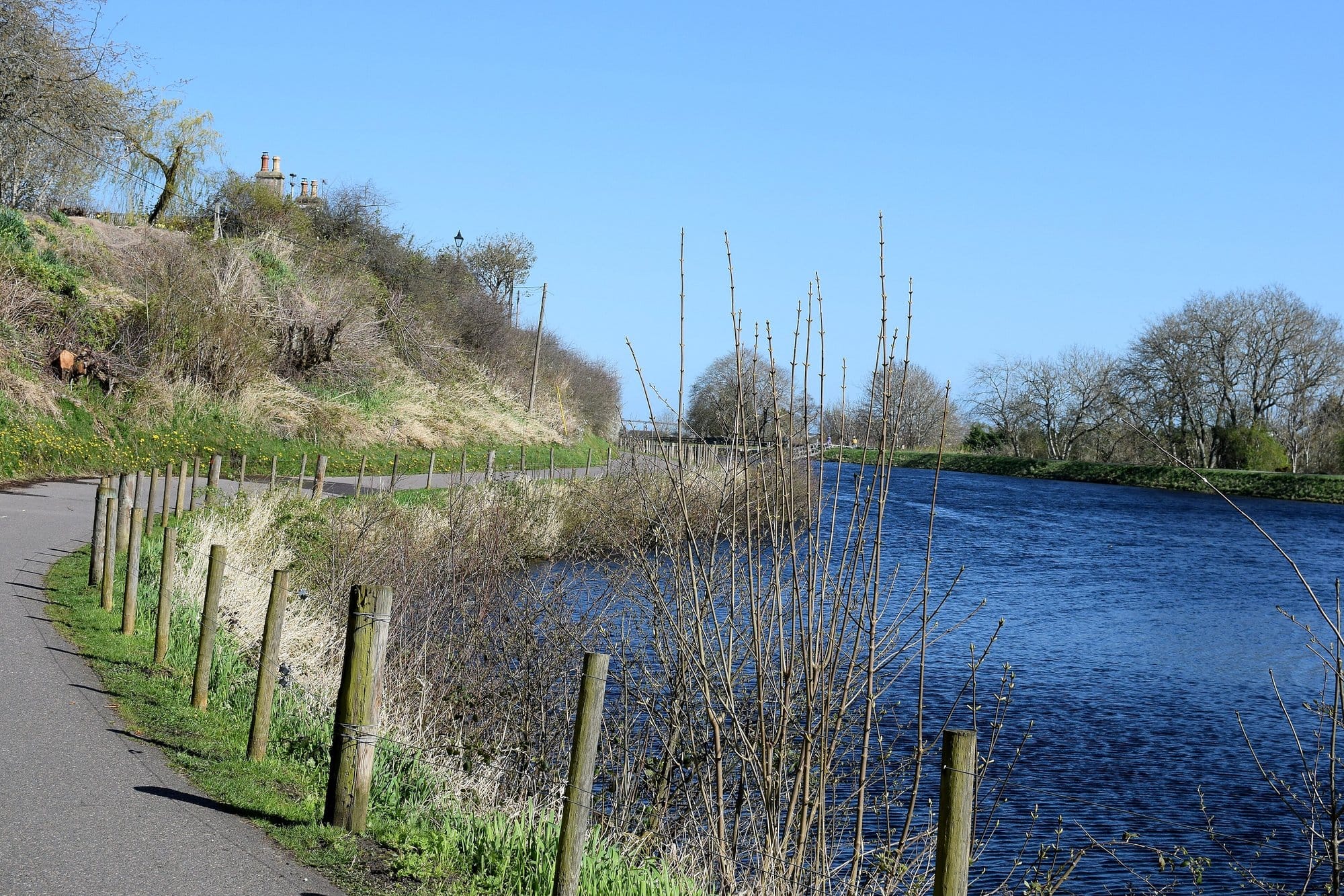 A paved towpath lined with wooden posts running beside the blue water of the Caledonian Canal under a clear sky