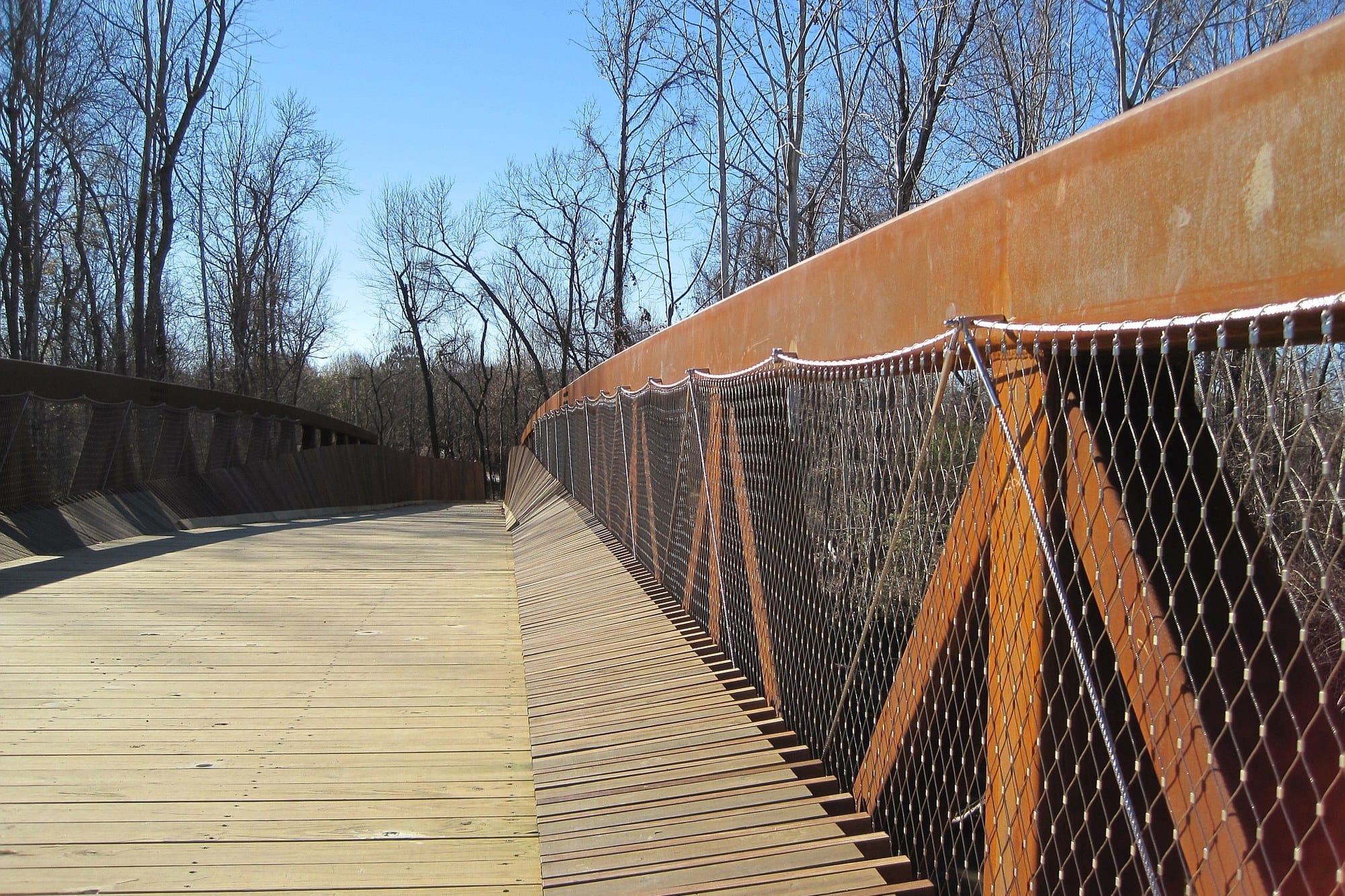 A timber boardwalk with a rusted steel rail and cable netting curving through bare winter woods on the Wolf River Greenway