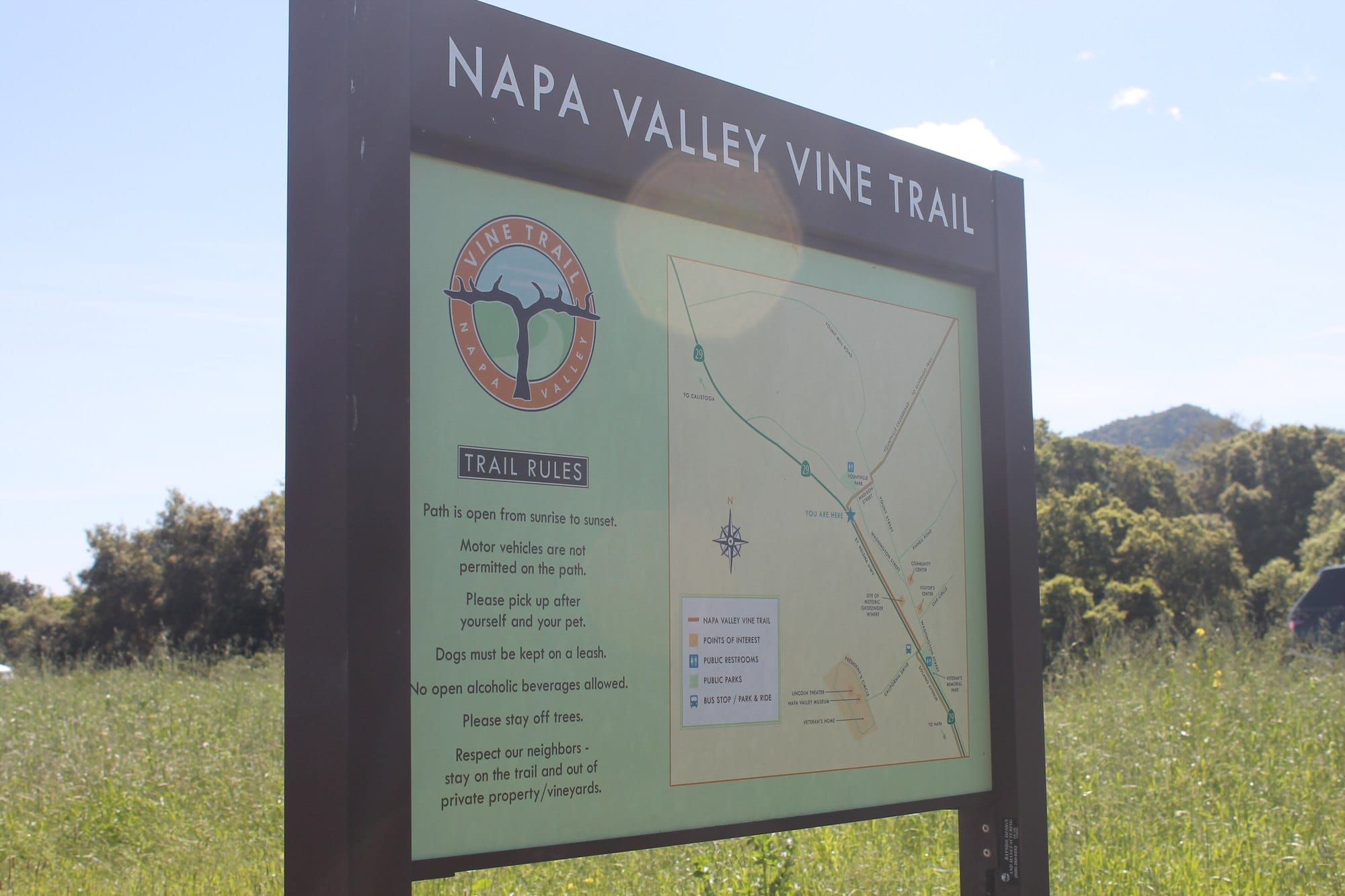 A Napa Valley Vine Trail sign with a map and trail rules, including that dogs must be kept on a leash, beside a grassy meadow