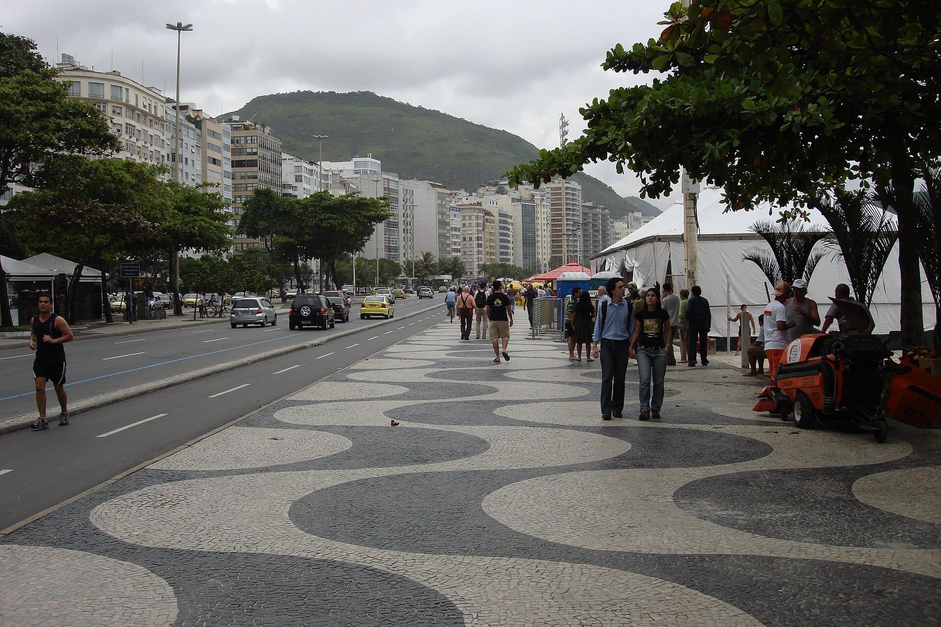 People walking on the black and white wave pavement of the Copacabana promenade under trees, with apartment blocks and a green hill behind