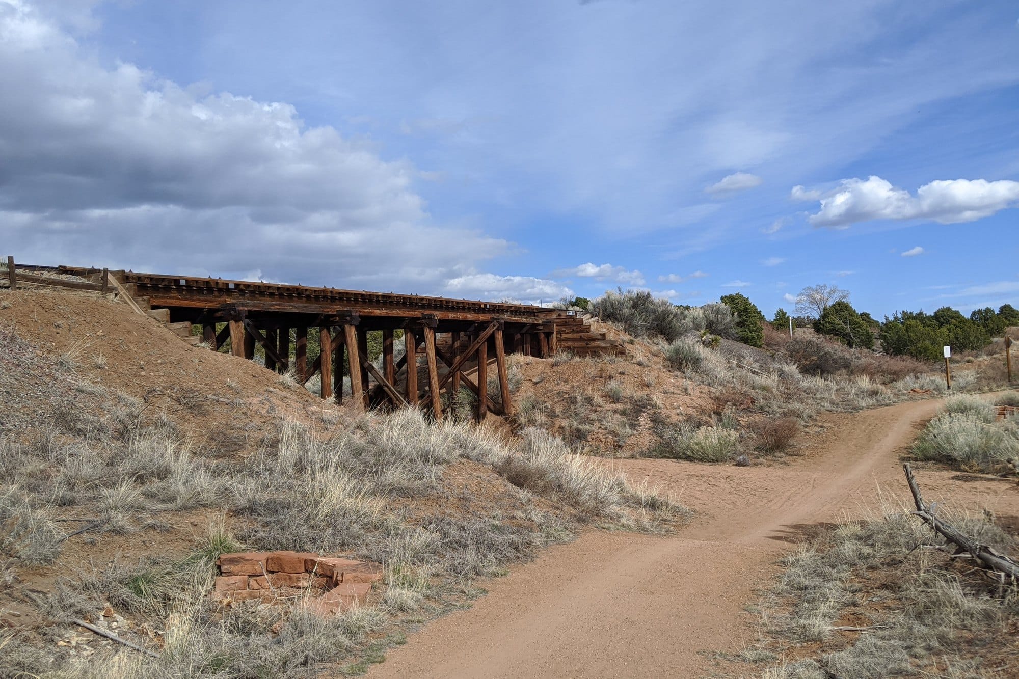 A dirt path curving past an old wooden railway trestle through desert scrub on the Santa Fe Rail Trail