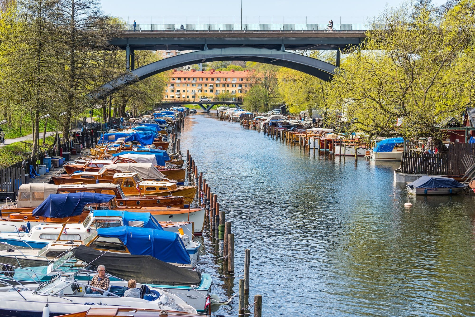 Wooden motorboats moored along a tree lined channel under a bridge at Långholmen, Stockholm, with a waterside path on the left