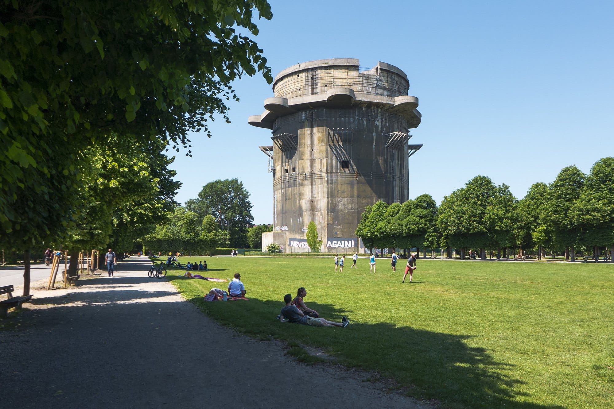 A shaded gravel path beside a wide lawn in the Augarten, with a concrete flak tower rising behind the trees