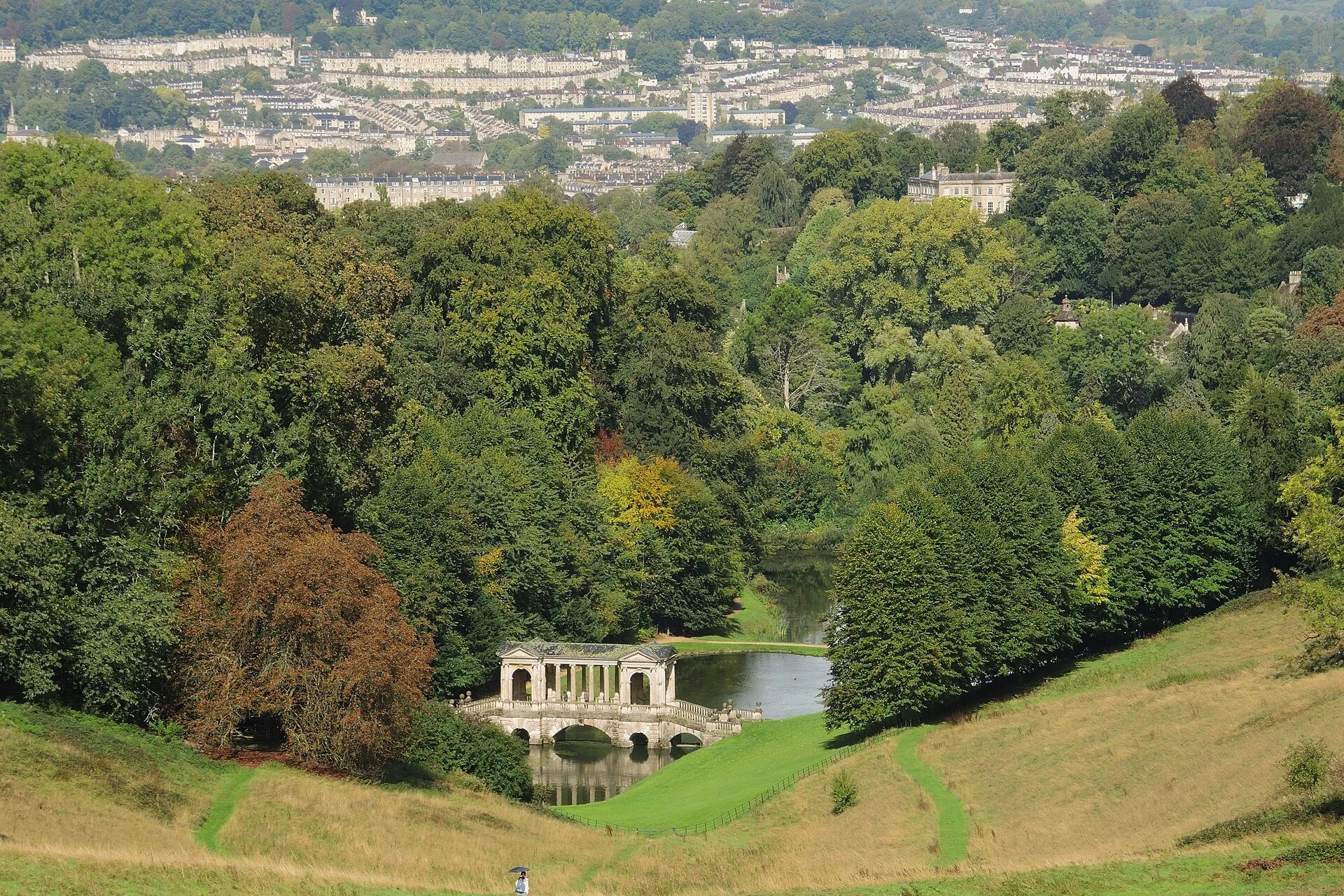 The Palladian Bridge and lake at Prior Park, with a path across the hillside and Bath's terraces beyond