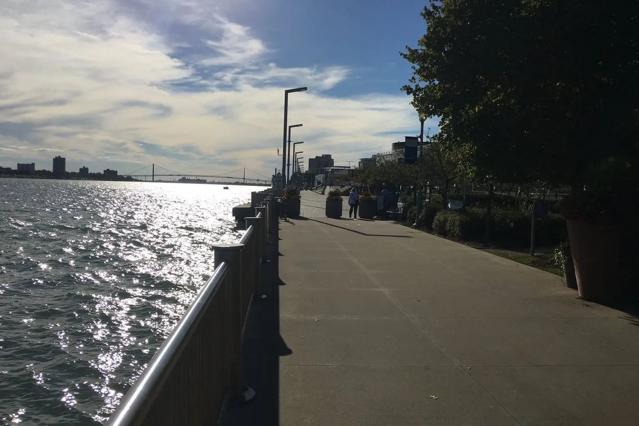The Detroit Riverwalk on a sunny afternoon, a railed promenade beside the sparkling Detroit River with a suspension bridge on the horizon