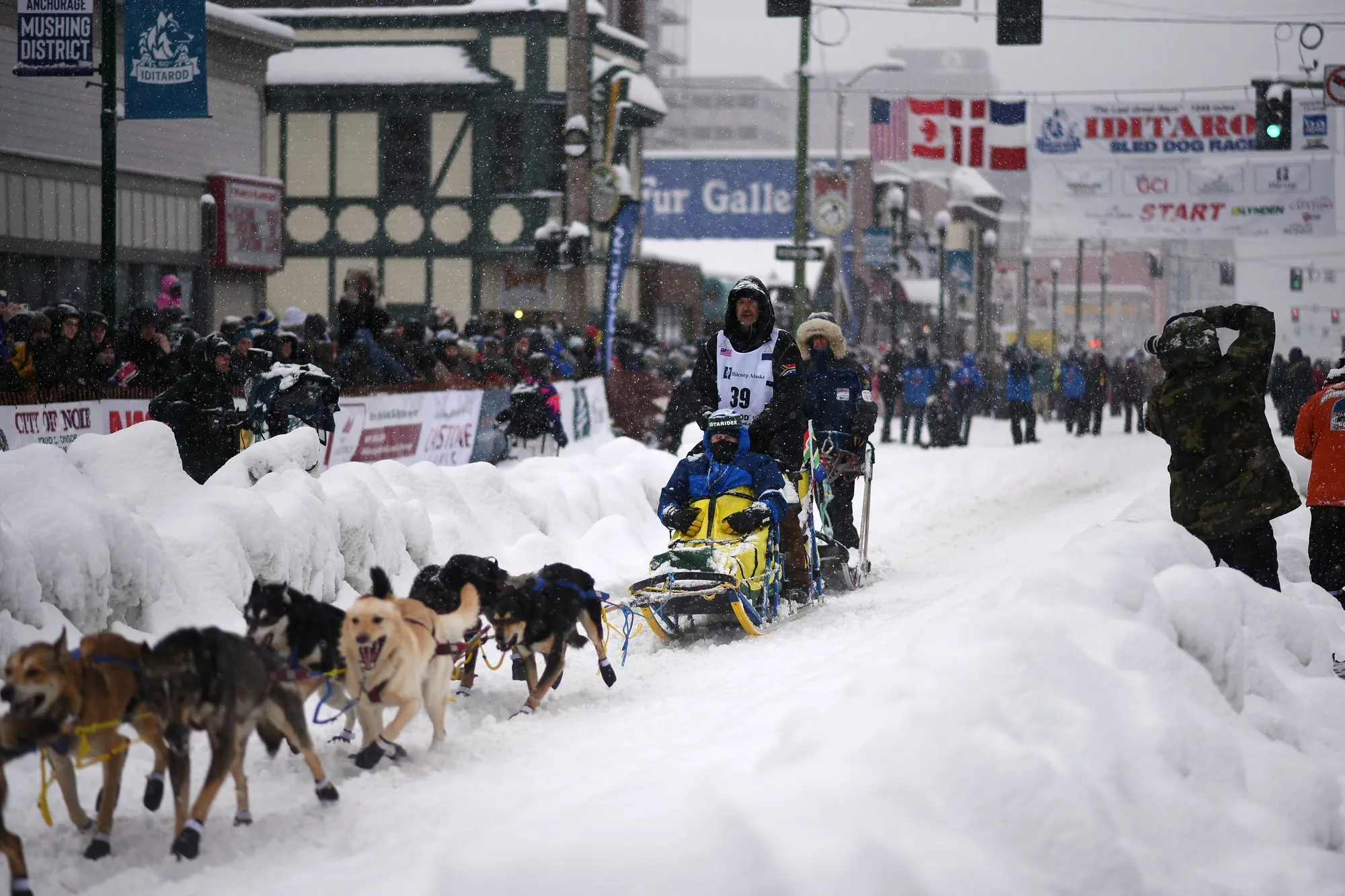 A musher and his sled dog team running down a snow covered Fourth Avenue in Anchorage past crowds and an Iditarod start banner
