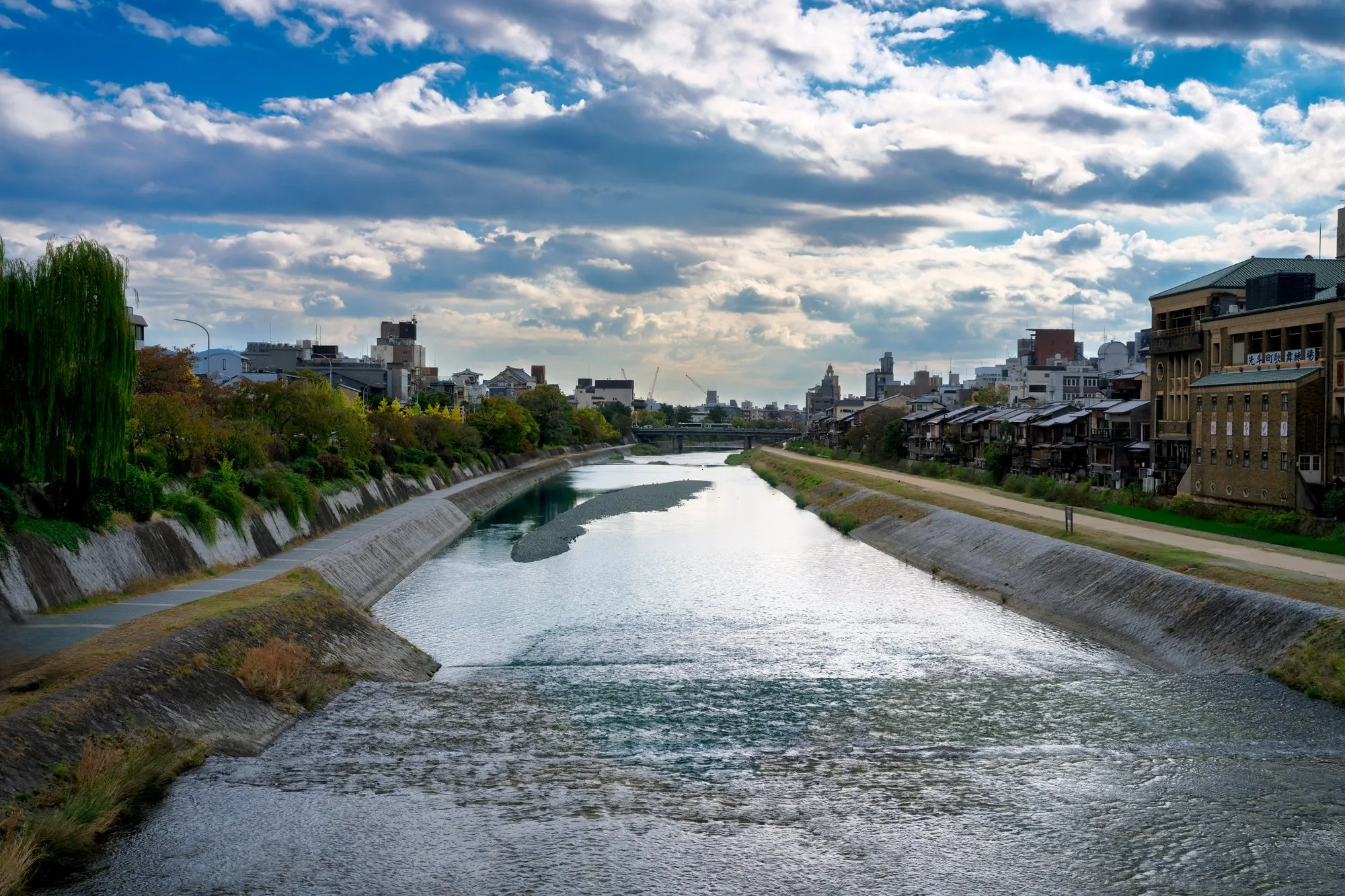 The Kamo River in Kyoto flowing between stone embankments, with walking paths along both banks and the city under a cloudy autumn sky