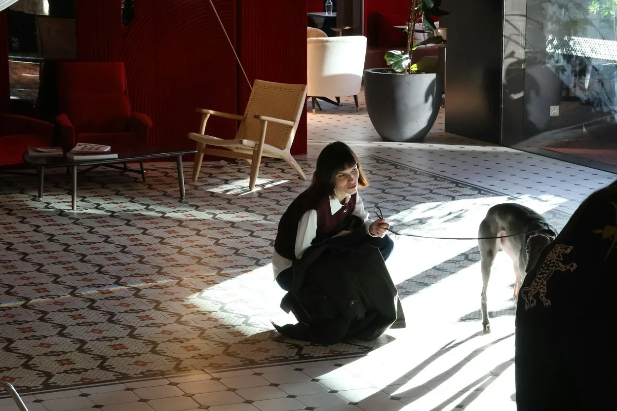 A woman crouching to greet a grey greyhound on a lead in a sunlit hotel lobby with a patterned tiled floor
