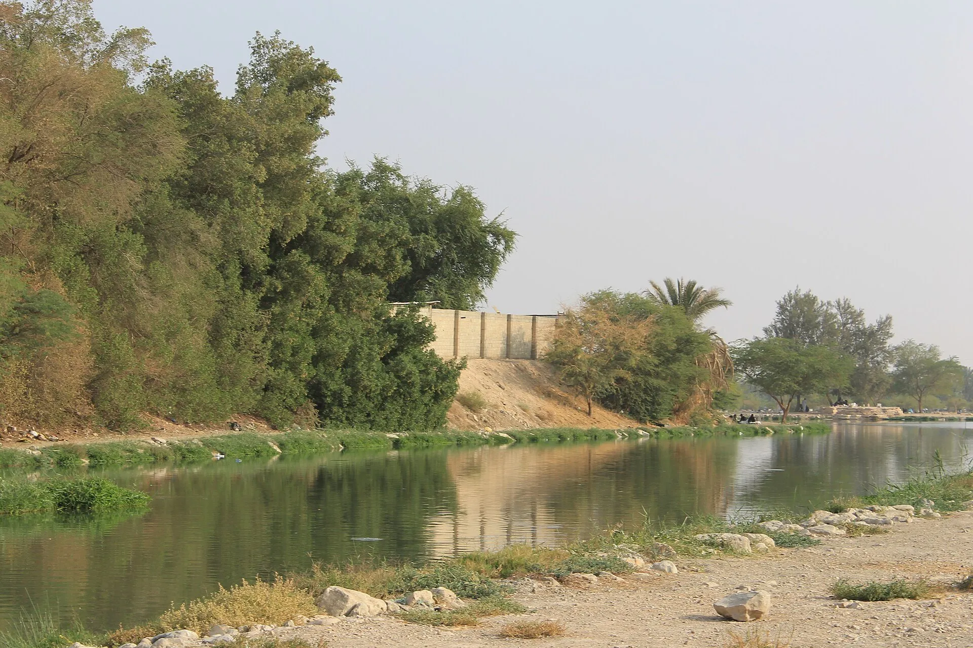 A still lake in Wadi Hanifah, Riyadh, with trees and palms along the far bank and a stony path along the near shore