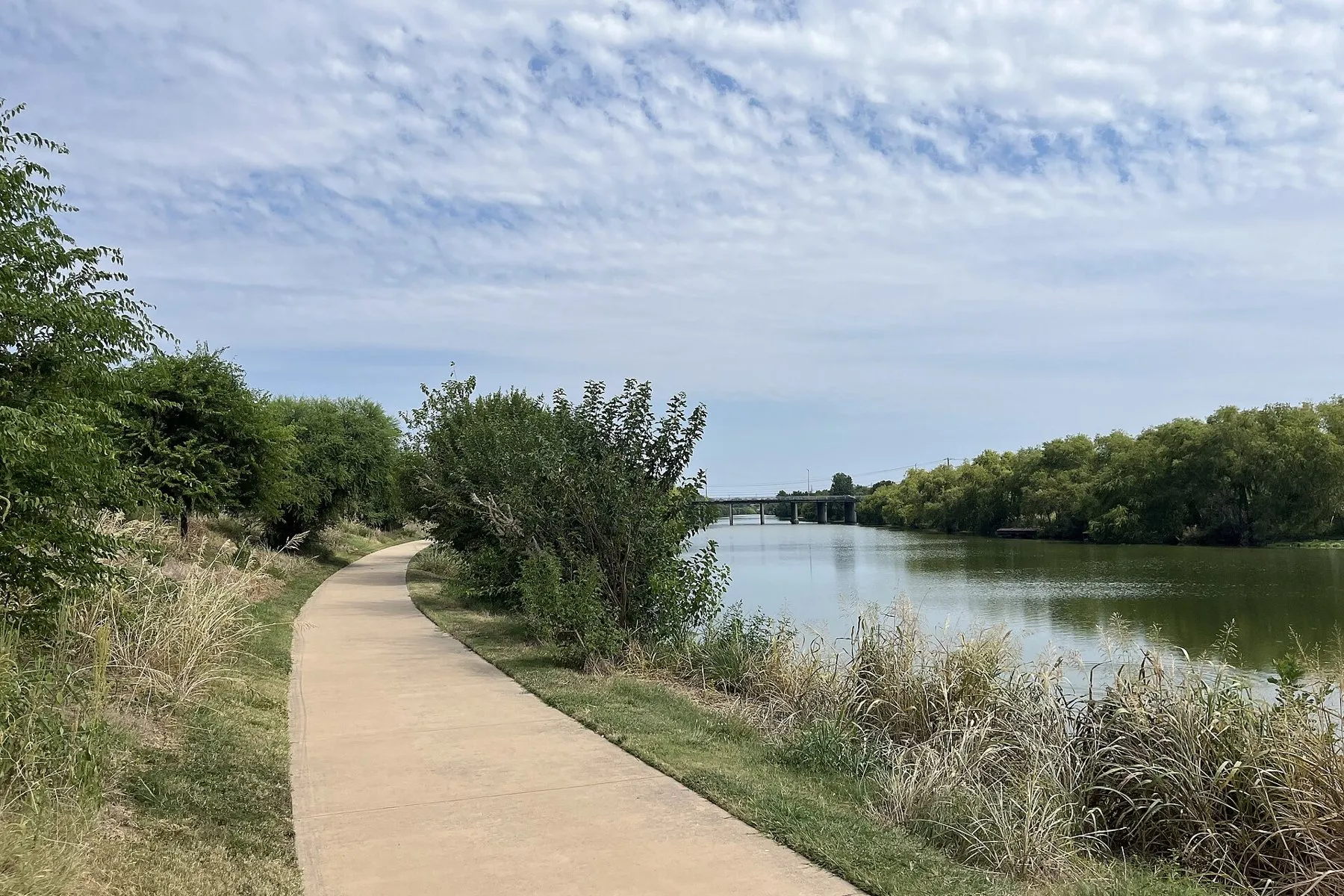 A paved riverside trail curving along the San Antonio River on the Mission Reach under a high cloudy sky
