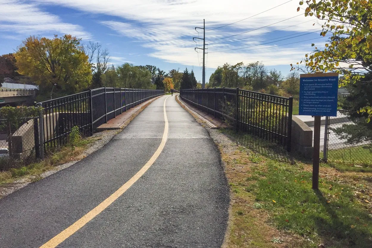 A paved path with a yellow centre line crossing a railed bridge on Grant's Trail in St. Louis, autumn trees on either side and a trail sign on the right