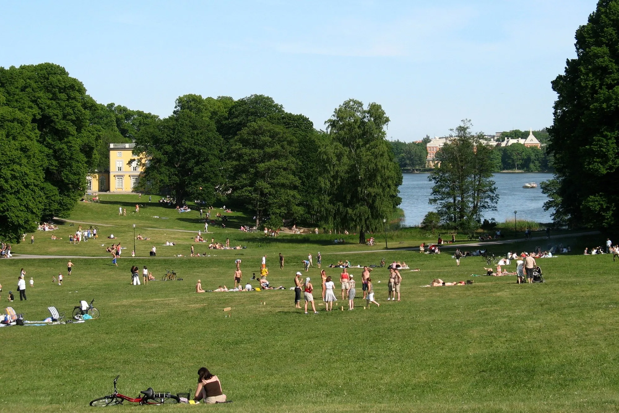 A wide lawn with people picnicking, a yellow pavilion among tall trees and the water of Brunnsviken beyond, in Haga Park, Stockholm