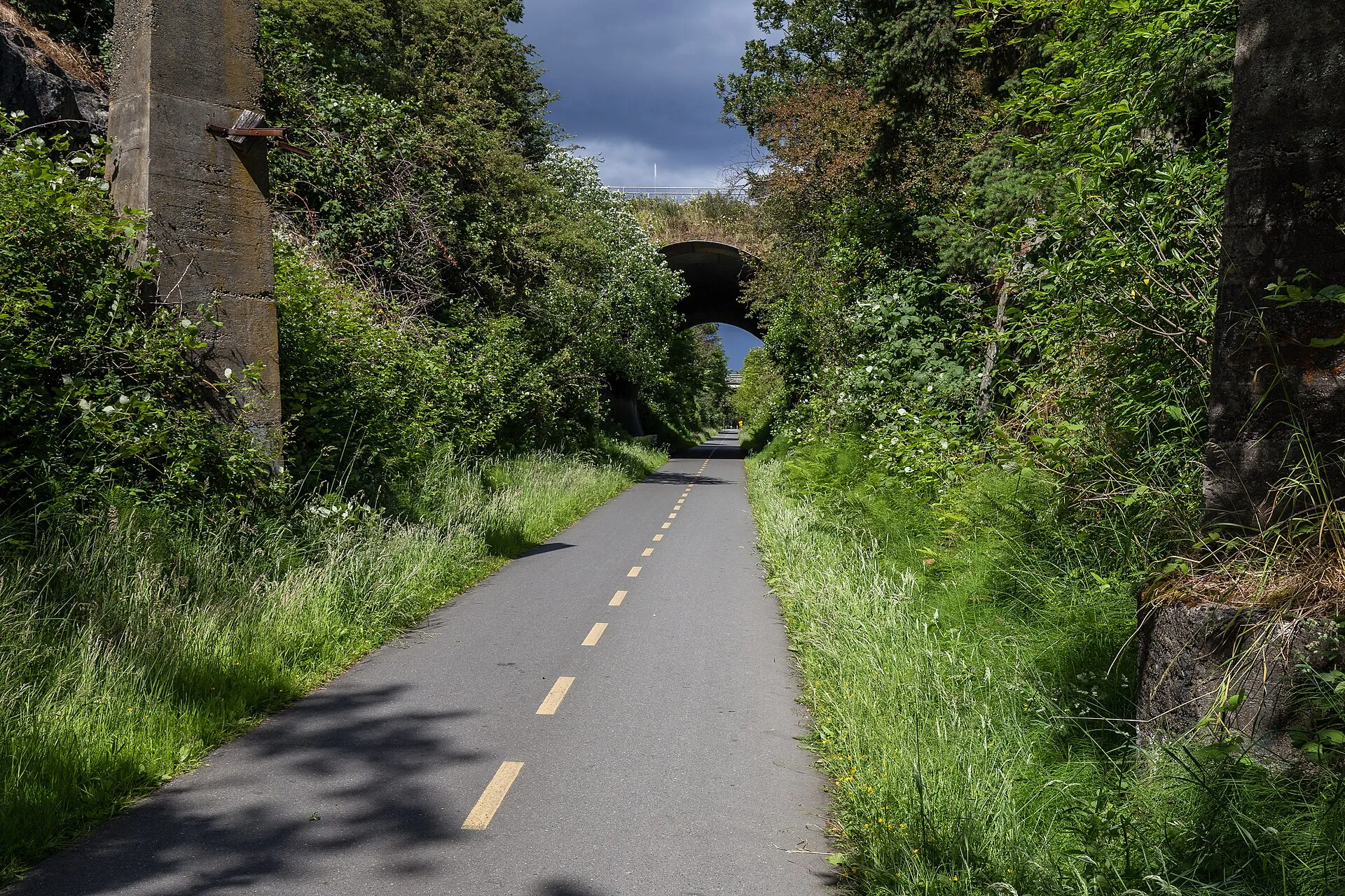 A paved section of the Galloping Goose Regional Trail running between green verges towards a stone bridge in Saanich