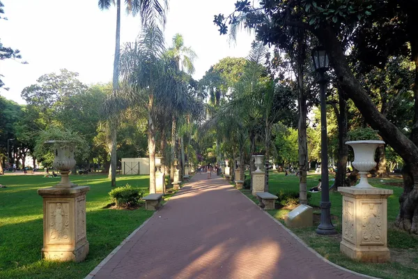 A brick path between stone urns, palms and old trees in Parque Lezama, San Telmo, with people walking in the distance