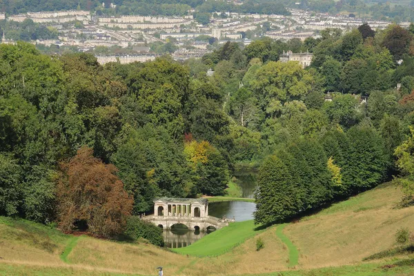The Palladian Bridge and lake at Prior Park, with a path across the hillside and Bath's terraces beyond