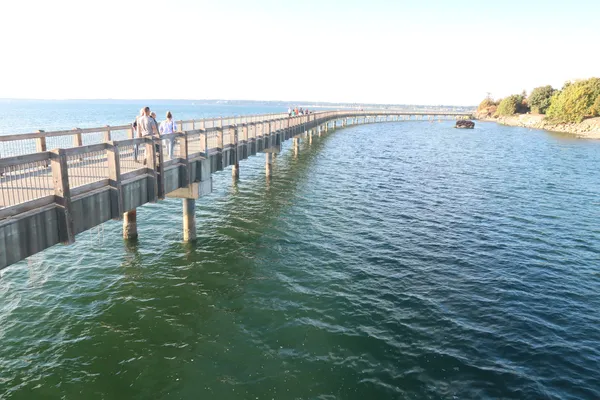 People walking along a curving wooden boardwalk over the blue water of Bellingham Bay, with wooded shoreline beyond