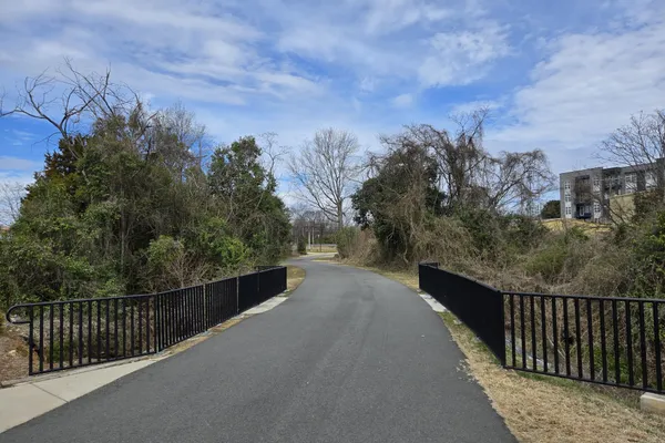 A paved greenway path crossing a bridge with black railings between trees and shrubs under a blue sky