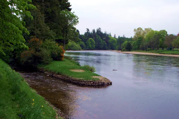 The River Ness flowing past grassy banks and tall trees at the Ness Islands in Inverness, with a stone edged inlet in the foreground