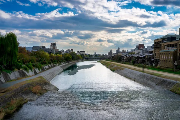 The Kamo River in Kyoto flowing between stone embankments, with walking paths along both banks and the city under a cloudy autumn sky