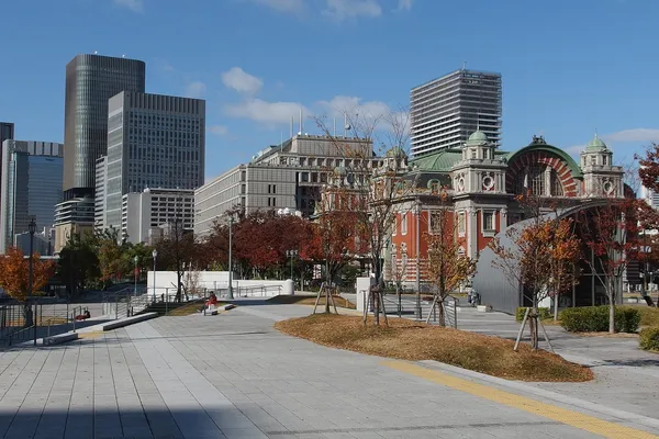 A wide paved riverside path in Nakanoshima Park, Osaka, with autumn trees and the red brick Central Public Hall