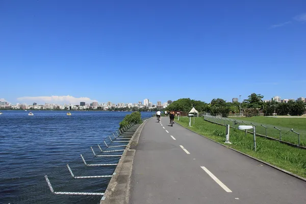 A cycle path and walkway running beside the Lagoa Rodrigo de Freitas, with grass on one side, the water on the other and Ipanema's buildings beyond