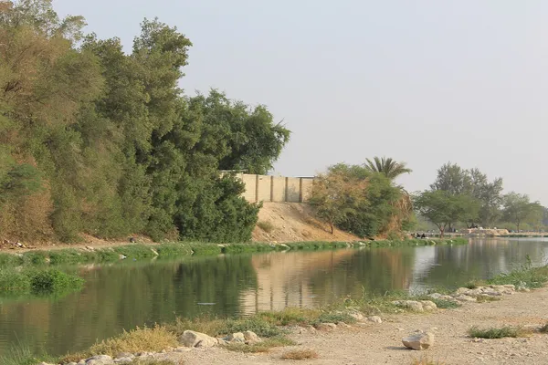 A still lake in Wadi Hanifah, Riyadh, with trees and palms along the far bank and a stony path along the near shore