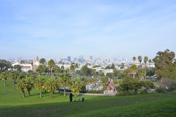 Green lawns and palm trees sloping down Mission Dolores Park towards the downtown San Francisco skyline under a blue sky