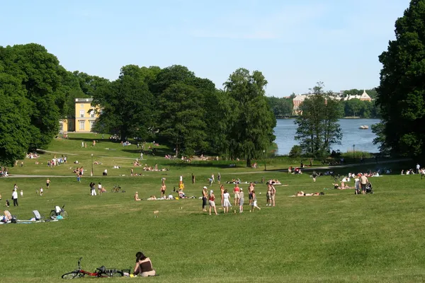 A wide lawn with people picnicking, a yellow pavilion among tall trees and the water of Brunnsviken beyond, in Haga Park, Stockholm